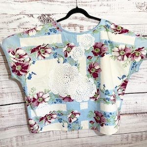 May include: A short-sleeved top featuring a light blue and white grid pattern with a floral print of dark pink and white flowers. The garment is embellished with several white crocheted doilies of varying sizes attached to the front center.