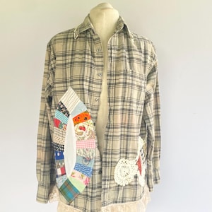 May include: A long-sleeve button-down shirt in a beige and black plaid pattern. The shirt features a large, oval-shaped patchwork applique on the right side, a white crocheted lace accent on the left, and a white lace trim along the bottom hem.
