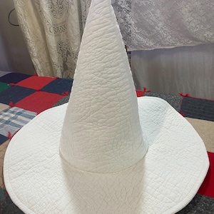 May include: A white pointed hat with a textured, quilted surface. The hat features a tall, conical crown and a wide, circular brim. The fabric has a distinct, repeating stitched pattern across the entire exterior, creating a subtle, raised, and crinkled appearance throughout the material.