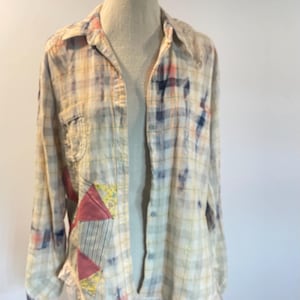 May include: A long-sleeve button-down shirt featuring a light-colored plaid pattern with blue and pink tie-dye accents. The shirt includes a patchwork design on the front with triangular fabric pieces and a white lace trim along the bottom hem.