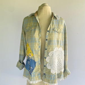 May include: A long-sleeve button-down shirt in a light blue and yellow plaid pattern. The shirt features a white lace trim on the collar and hem, a large white circular crochet doily on the front, and two fabric patches in yellow and blue.
