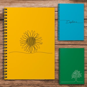 May include: Three spiral-bound notebooks on a wooden surface. A yellow notebook features a black line drawing of a sunflower. A blue notebook has the word "Explore" in script. A green notebook displays a tree outline.