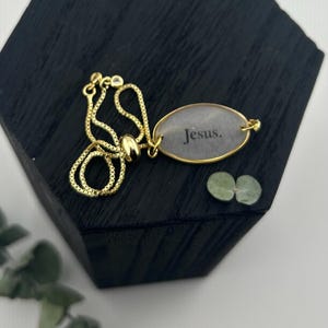 May include: A gold-colored adjustable bracelet with an oval pendant. The pendant has the word "Jesus" in black lettering. The bracelet is on a black wooden block with a white background.