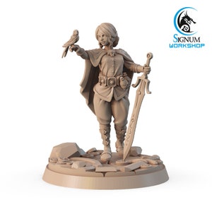 May include: A miniature figurine of a female character with short blonde hair, wearing a brown tunic and pants, holding a sword and a small bird. The figurine is standing on a circular base with a textured surface.