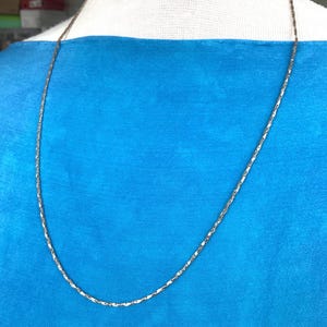Peut inclure: Un collier chaîne en métal argenté fin avec un motif de maillons texturés et répétitifs. Le collier est présenté sur un fond en tissu bleu vif, mettant en valeur l'éclat métallique et la surface complexe et réfléchissante des maillons.