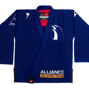 Op de afbeelding: Een marineblauwe Jiu-Jitsu gi met witte logo's en tekst. De gi heeft een wikkelontwerp met een rode binnenkraag. De logo's omvatten een bliksemschicht en een gestileerde vogel. De tekst luidt "ALLIANCE JIU JITSU TEAM".