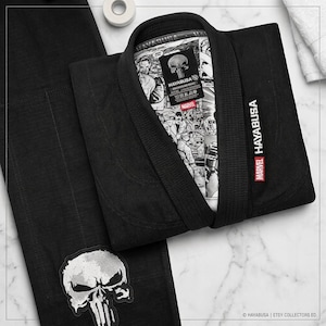 May include: A black martial arts gi featuring a Marvel Punisher graphic on the inner lining and a large white Punisher skull patch on the leg. The collar displays Hayabusa and Marvel branding, with a comic book style print inside the jacket.