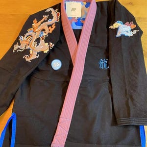 Op de afbeelding: Zwarte judojas met een roze band. Het uniform heeft een gedetailleerde geborduurde draak op de linkermouw en een blauw-witte patch op de rechtermouw. Het woord "龍" is blauw op de borst geborduurd.