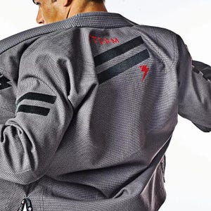 Pode incluir: Kimono de Jiu-Jitsu cinza com listras pretas e um logotipo "TEAM" vermelho nas costas.