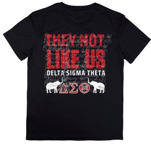 Puede incluir: Camiseta negra con el texto "THEY NOT US" en rojo y blanco. Debajo, "DELTA SIGMA THETA" en blanco. La camiseta presenta dos gráficos de elefantes blancos y las letras griegas ΔΣΘ.