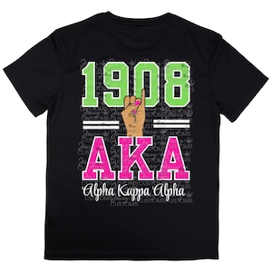 May include: Black t-shirt with the text "1908" in green, a hand gesture, and "AKA" in pink. The words "Alpha Kappa Alpha" are written below in white script. The shirt has a crew neck.