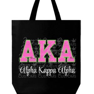 May include: Black tote bag with the letters "AKA" in pink with a green outline. The words "Alpha Kappa Alpha" are written in white script below the letters. The background has a repeating pattern of the word "CUSTOMS".