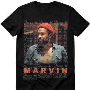 May include: Black t-shirt featuring a portrait of a man wearing a red beanie and denim shirt. The name "MARVIN" is printed in orange block letters. The background has a repeating pattern of "Jazzy Customs".