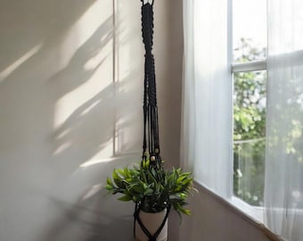 May include: A black macrame plant hanger with a white ceramic pot holding a green plant. The plant hanger is suspended from a hook and features wooden beads. The plant is positioned near a window with sheer curtains.