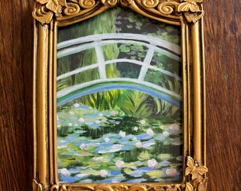 May include: A gold-colored ornate frame with a butterfly design on the top and sides. The frame holds a painting of a bridge over a pond with water lilies.