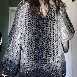 May include: A gray, open-weave crochet cardigan with a gradient effect, transitioning from light gray to a deep charcoal at the hem. The sleeves are wide and the overall design is loose-fitting.