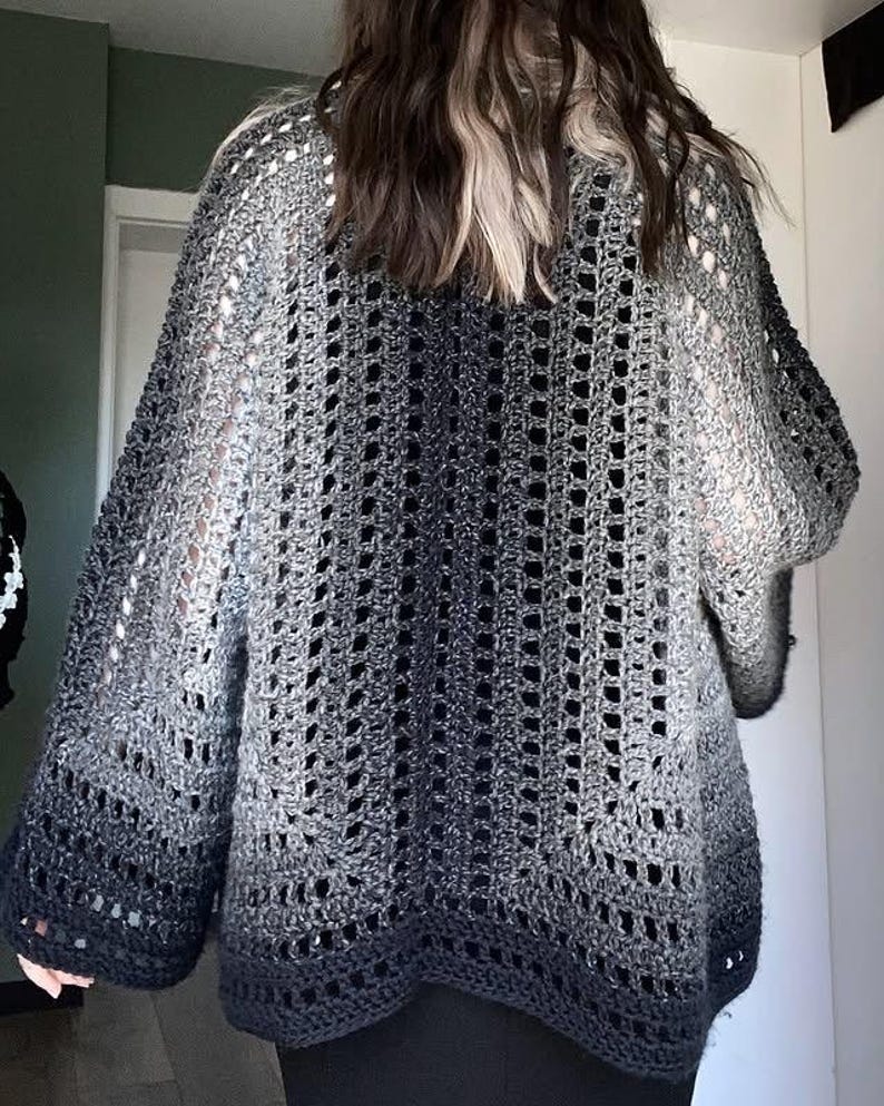 May include: A gray, open-weave crochet cardigan with a gradient effect, transitioning from light gray to a deep charcoal at the hem. The sleeves are wide and the overall design is loose-fitting.