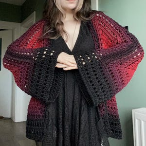 May include: A handmade crochet cardigan in a gradient of red, purple, and black. The open-front design features wide, bell sleeves with a lacy pattern. The cardigan is worn over a black dress, creating a layered look.