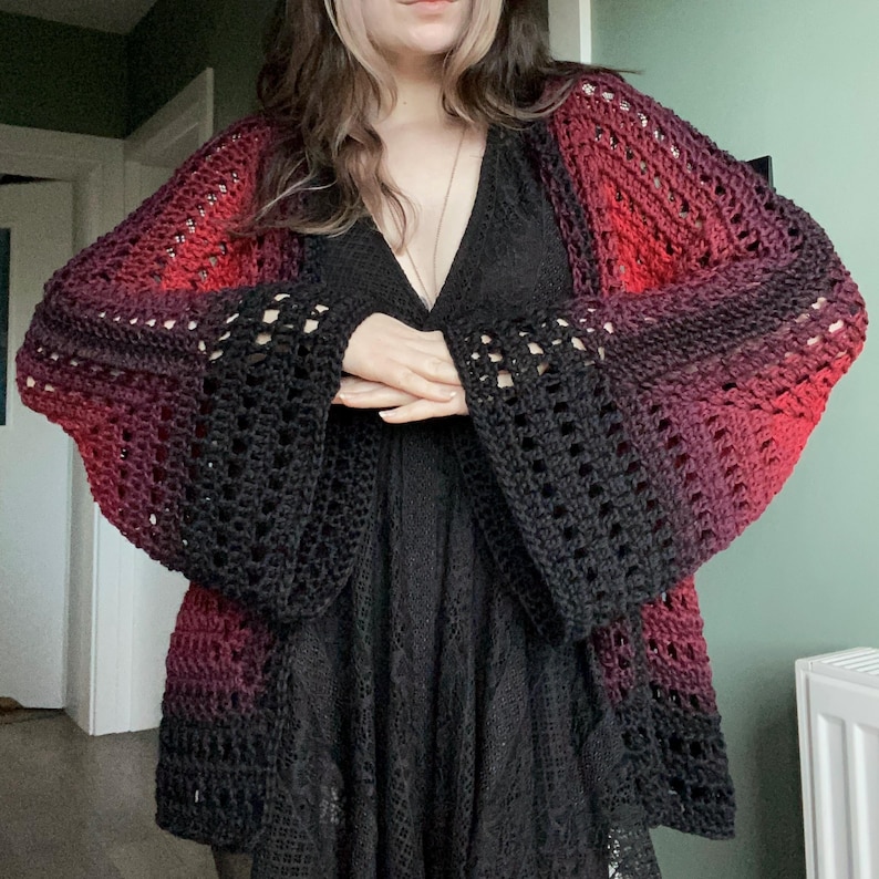 May include: A handmade crochet cardigan in a gradient of red, purple, and black. The open-front design features wide, bell sleeves with a lacy pattern. The cardigan is worn over a black dress, creating a layered look.