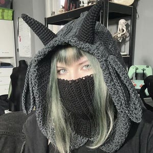 May include: A dark gray crocheted hood with pointed ears and a face covering. The hood is made of a thick yarn and has a textured appearance. The face covering is also crocheted and covers the lower face.