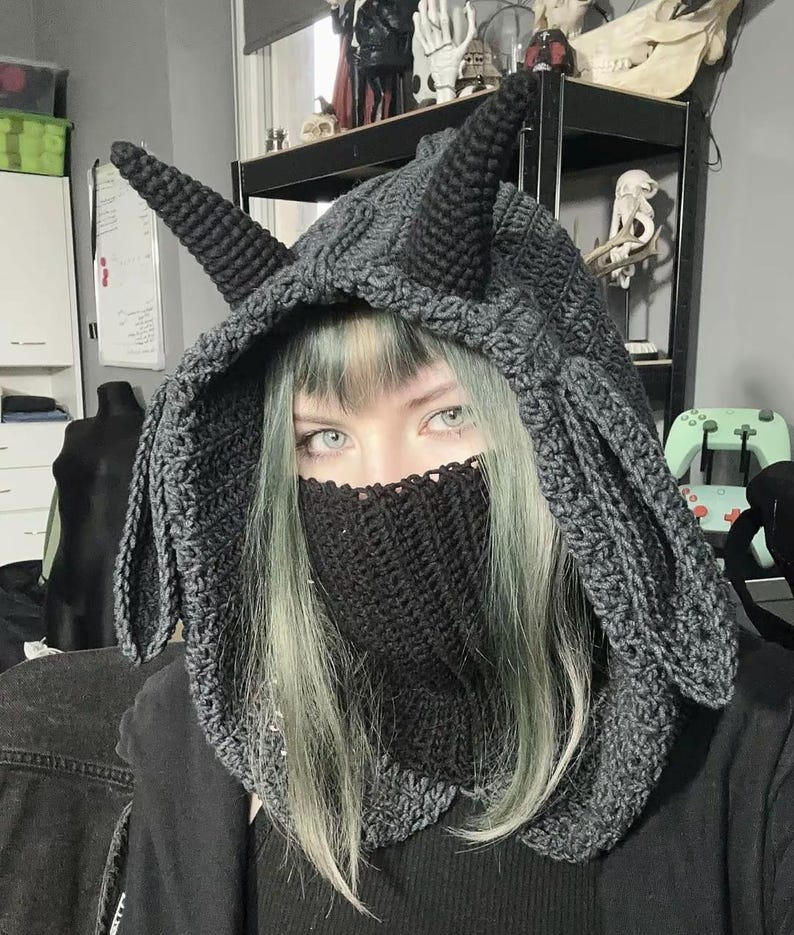 May include: A dark gray crocheted hood with pointed ears and a face covering. The hood is made of a thick yarn and has a textured appearance. The face covering is also crocheted and covers the lower face.