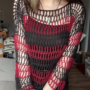 May include: A black and red striped open-weave sweater. The sweater has a round neckline and long sleeves. The red stripes alternate with black stripes. A black vest is visible underneath the sweater. A silver pendant hangs from a black necklace.