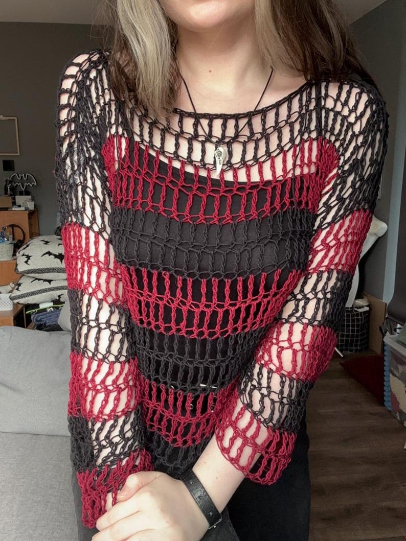 May include: A black and red striped open-weave sweater. The sweater has a round neckline and long sleeves. The red stripes alternate with black stripes. A black vest is visible underneath the sweater. A silver pendant hangs from a black necklace.