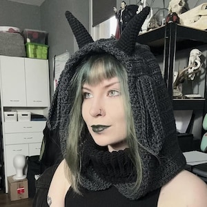 May include: A dark gray crocheted hood with two pointed horns. The hood is designed to cover the head and neck, with a cowl-like design. The hood is made of a thick yarn, and the horns are also crocheted. The hood is a unique accessory.