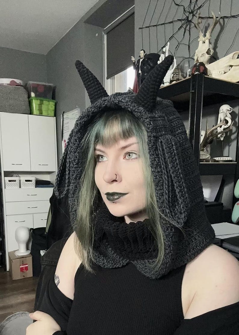 May include: A dark gray crocheted hood with two pointed horns. The hood is designed to cover the head and neck, with a cowl-like design. The hood is made of a thick yarn, and the horns are also crocheted. The hood is a unique accessory.