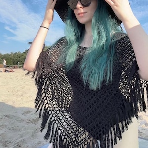May include: A black crochet poncho featuring an open-knit pattern and long fringe trim along the edges. The garment is worn with a wide-brimmed black sun hat and dark-tinted sunglasses, creating a layered look with a textured, woven appearance.