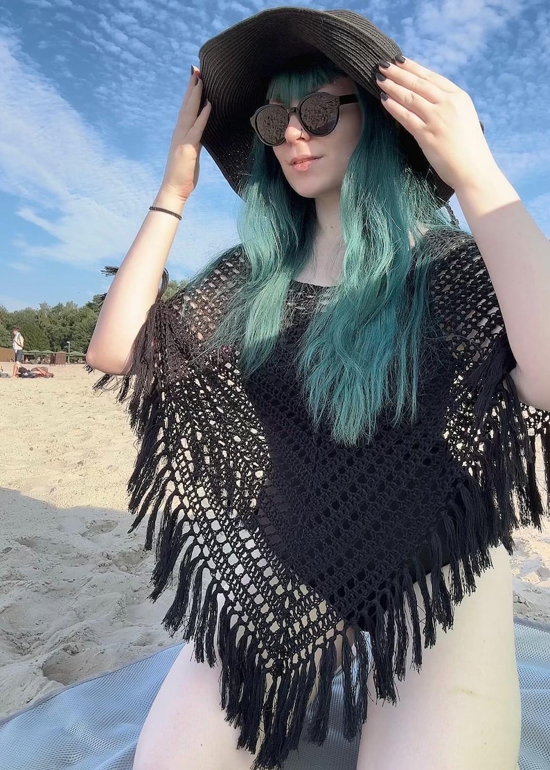 May include: A black crochet poncho featuring an open-knit pattern and long fringe trim along the edges. The garment is worn with a wide-brimmed black sun hat and dark-tinted sunglasses, creating a layered look with a textured, woven appearance.