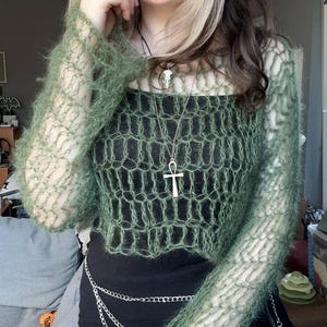 May include: A green, open-weave, long-sleeved top with a matching cropped sweater. The outfit includes a black top, a silver chain belt, and a silver Ankh pendant necklace. The style is bohemian and alternative.