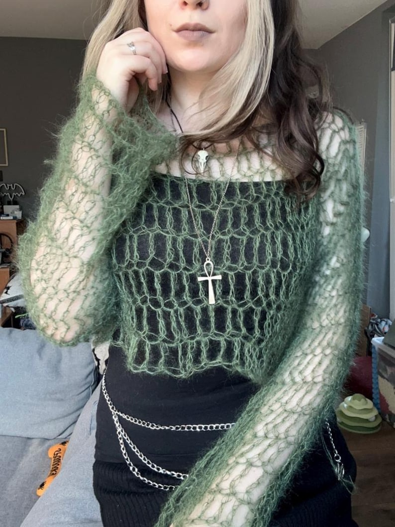 May include: A green, open-weave, long-sleeved top with a matching cropped sweater. The outfit includes a black top, a silver chain belt, and a silver Ankh pendant necklace. The style is bohemian and alternative.
