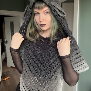 May include: A dark gray crochet hooded poncho featuring two prominent, pointed horns on the hood. The garment displays an ombre color transition from a deep charcoal at the top to a lighter gray at the bottom, with a V-shaped hem and openwork.
