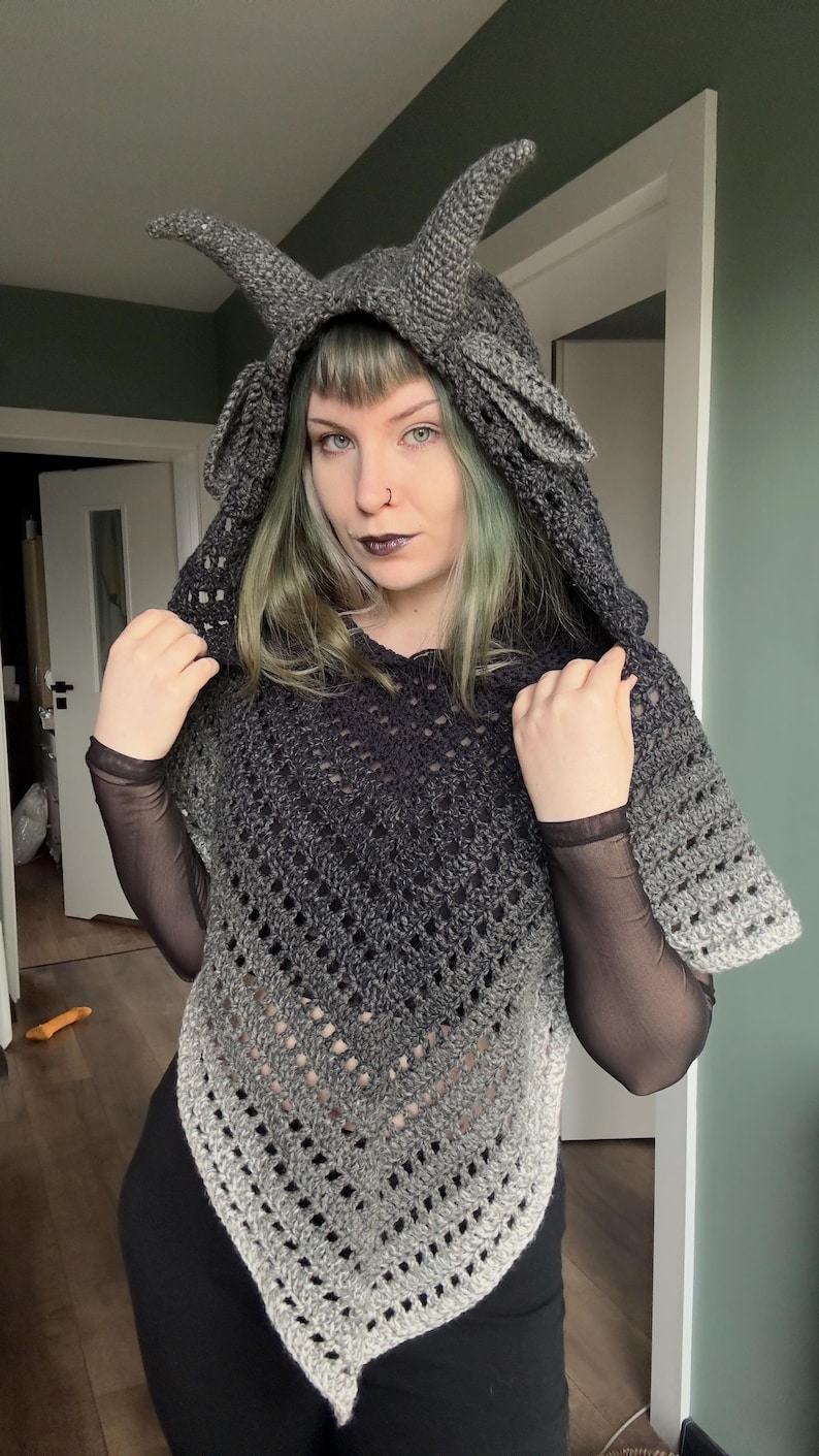 May include: A dark gray crochet hooded poncho featuring two prominent, pointed horns on the hood. The garment displays an ombre color transition from a deep charcoal at the top to a lighter gray at the bottom, with a V-shaped hem and openwork.