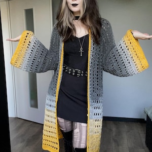 May include: A long, open-front crochet cardigan in shades of gray and yellow. The cardigan features wide sleeves with a yellow ombre effect at the cuffs and a yellow trim along the edges. The open weave design adds a light and airy feel to the garment.