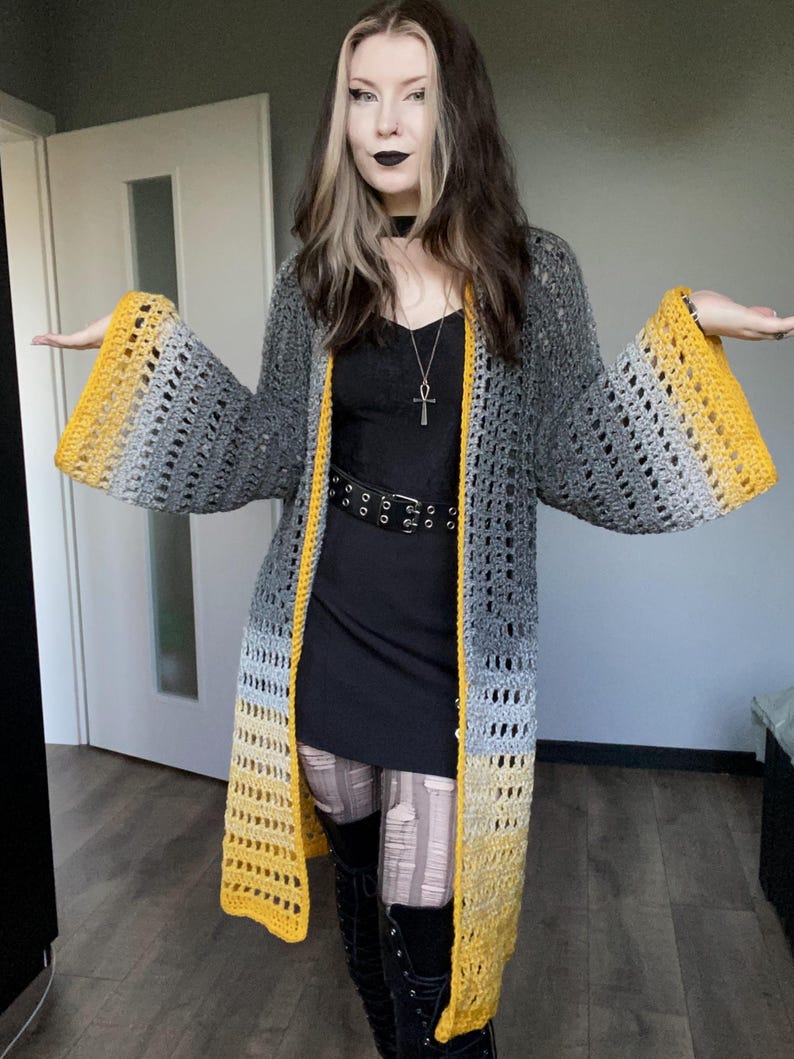 May include: A long, open-front crochet cardigan in shades of gray and yellow. The cardigan features wide sleeves with a yellow ombre effect at the cuffs and a yellow trim along the edges. The open weave design adds a light and airy feel to the garment.