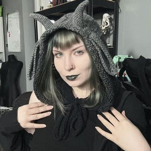 May include: A gray crocheted hood with pointed horns and long, draped sides. The hood is tied at the neck with a black crocheted tie. The hood is a unique accessory.