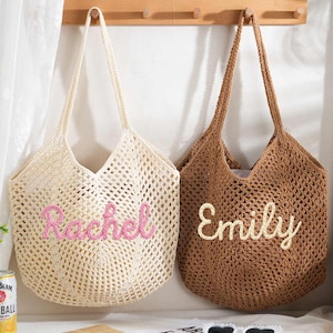 Op de afbeelding: Twee gehaakte draagtassen, één crème en één bruin, hangen aan een houten rek. De crèmekleurige tas heeft de naam "Rachel" in roze, en de bruine tas heeft "Emily" in crème. De tassen zijn open geweven.
