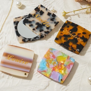 May include: Four square compact mirrors with personalized names engraved in gold script. One mirror is open, revealing a circular glass surface. The cases feature various patterns including tortoiseshell, striped, and multicolored acrylic designs.