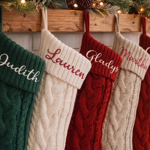 Puede incluir: Cinco calcetines navideños de punto colgados en fila, con patrones de trenzas y puños doblados. Los calcetines son de color verde oscuro, crema, rojo, beige y rojo. Cada puño está bordado con los nombres Judith, Lauren, Gladys, Martha y Crystal.