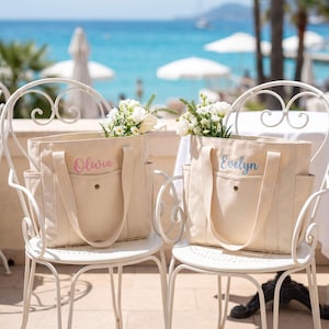 Pode incluir: Duas bolsas de lona em tom off-white com bolsos laterais e fecho de botão de pressão frontal. Uma bolsa apresenta o nome Olivia em escrita rosa, e a outra o nome Evelyn em escrita azul. Ambas as bolsas estão preenchidas com flores brancas.