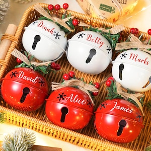May include: A selection of Christmas bell decorations in a wicker basket. The bells are red and white, with names such as "Betty" and "Marks" inscribed. Each bell features a keyhole design and is adorned with festive greenery and berries.
