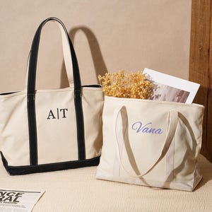 May include: Two canvas tote bags. One is cream-colored with black trim and handles, embroidered with "A|T". The other is cream-colored with the name "Vana" embroidered in blue. A coffee cup and newspaper are also visible.