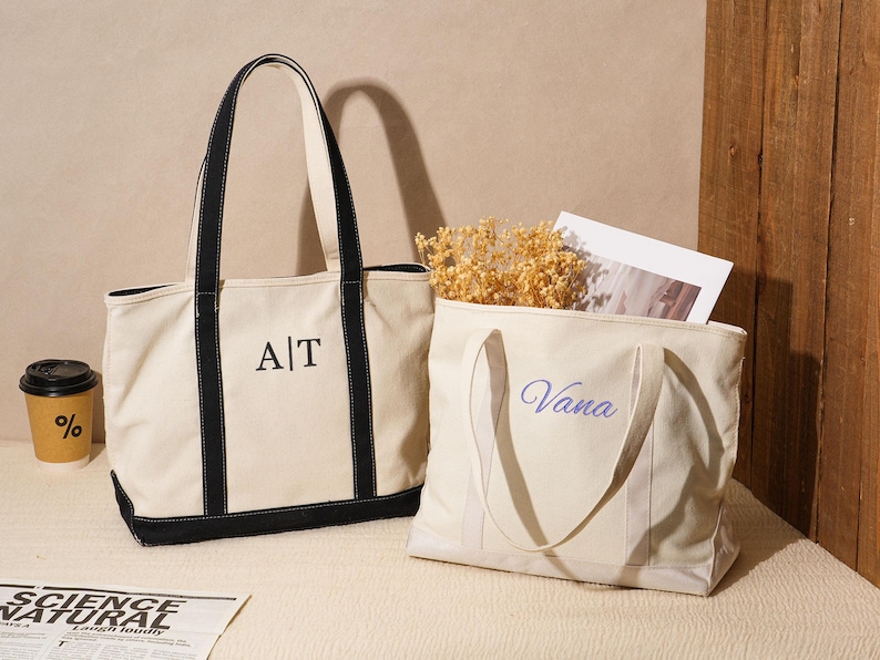 May include: Two canvas tote bags. One is cream-colored with black trim and handles, embroidered with "A|T". The other is cream-colored with the name "Vana" embroidered in blue. A coffee cup and newspaper are also visible.
