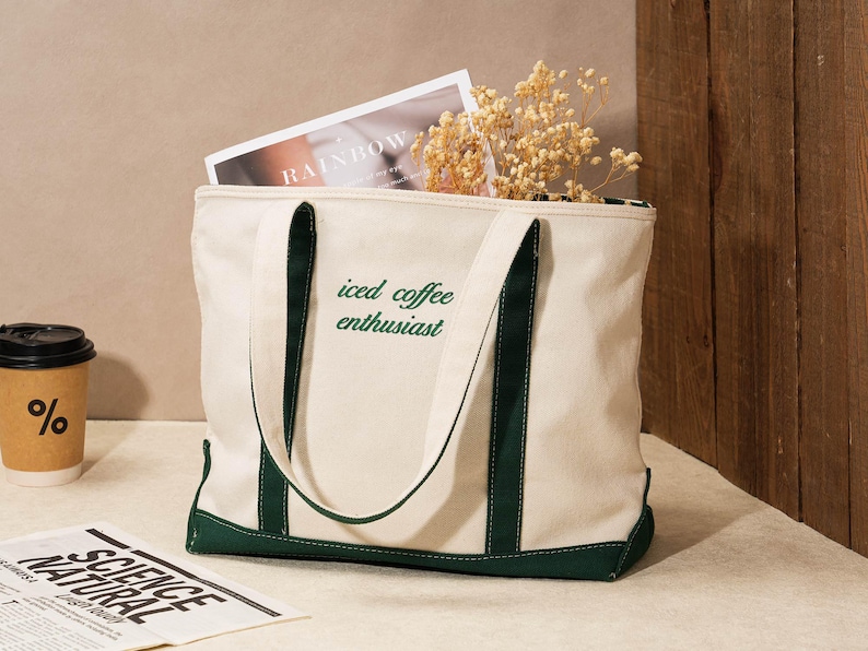 May include: Cream-colored tote bag with dark green accents and handles. The bag features the text "iced coffee enthusiast" in green. Inside the bag are a magazine and dried flowers. A coffee cup and newspaper are nearby.