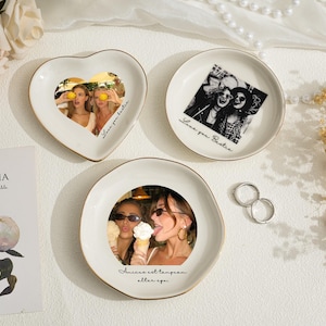 May include: Three white ceramic trinket dishes with gold rims, each featuring a photograph. One is heart-shaped, the others round. The photos depict people. Text on the dishes reads "Love you bestie" and "Amicae est temporam alter ego."
