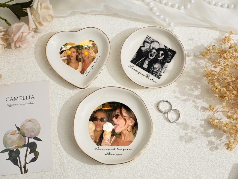 May include: Three white ceramic trinket dishes with gold rims, each featuring a photograph. One is heart-shaped, the others round. The photos depict people. Text on the dishes reads "Love you bestie" and "Amicae est temporam alter ego."