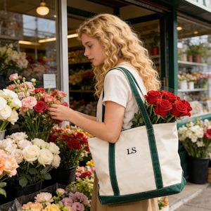 May include: A woman at a flower shop selects flowers, holding a canvas tote bag filled with red roses. The bag is off-white with green trim and the initials "LS". The shop displays a variety of colorful flowers.