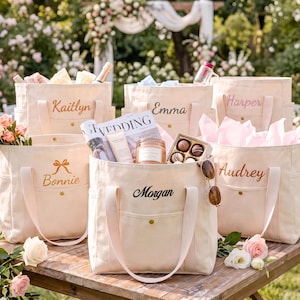 May include: A collection of six cream-coloured canvas tote bags, each featuring a front pocket with a gold snap button and personalised embroidery. The names Kaitlyn, Emma, Harper, Bonnie, Morgan, and Audrey are stitched on the front of the bags.